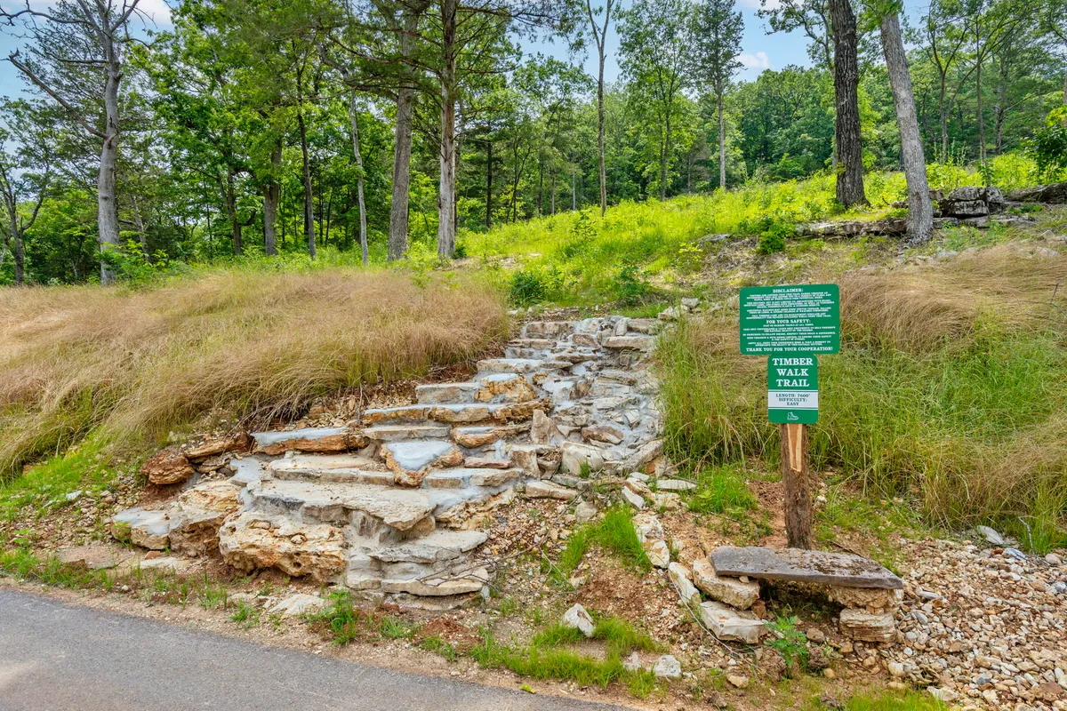 A rustic stone trail sign marks the beginning of a scenic woodland walking path, surrounded by abundant greenery and dappled sunlight. The inviting trail suggests peaceful strolls and opportunities for exploration and connection with nature.