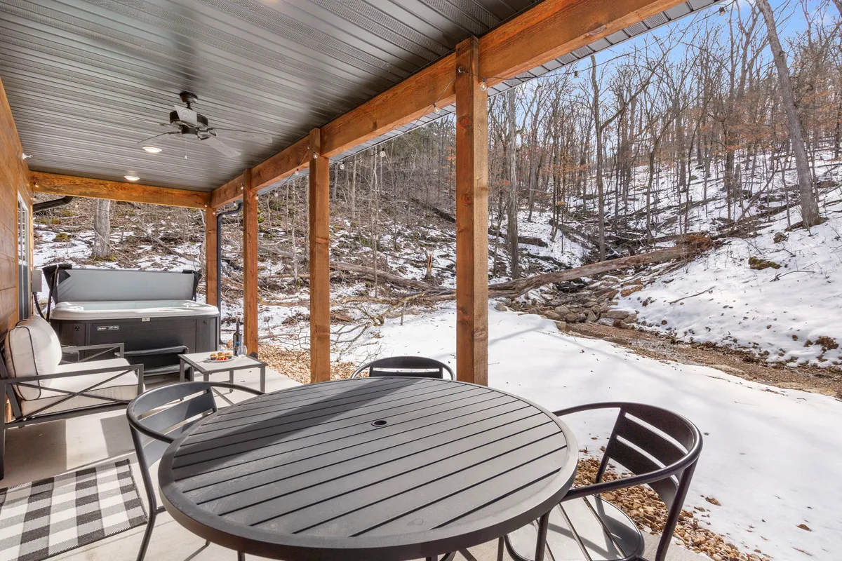 A tranquil outdoor area with a private hot tub set on a deck, surrounded by snow-covered landscape and a tree line for privacy. An outdoor dining table nearby offers opportunities for relaxation under the stars.