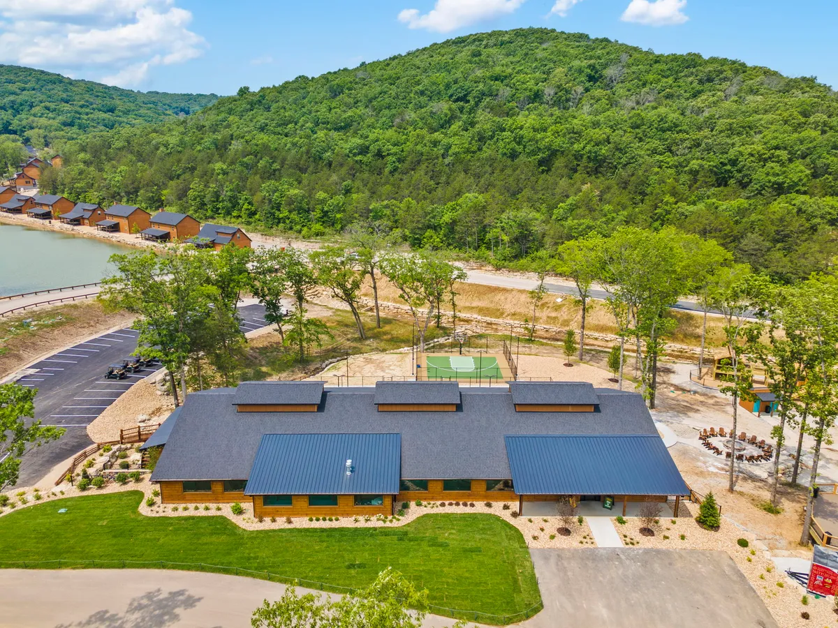 The main building and several cabins appear in the background, embraced by a lush tree line and an inviting lake. Recreational features such as a basketball court and open spaces suggest ample opportunities for activities and social gatherings. The scene has a relaxed, communal vibe, perfect for group adventures or solo relaxation.