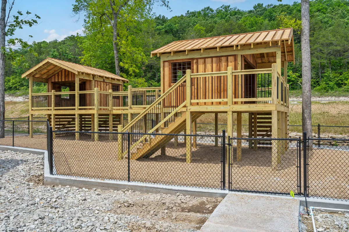 This image showcases a fenced outdoor play area with wooden playhouses and stairs, surrounded by natural greenery. Children are pictured exploring and playing, highlighting imaginative and active fun in a safe, inviting environment.
