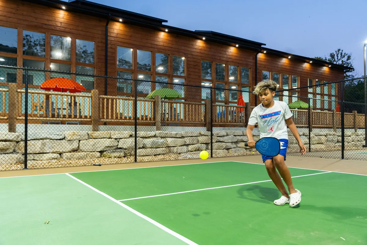A playful pickleball scene outside, with a young boy mid-game and friends nearby, set against the backdrop of a wooden building and colorful umbrellas. The setting is energetic and inviting, emphasizing friendly competition and outdoor activity.