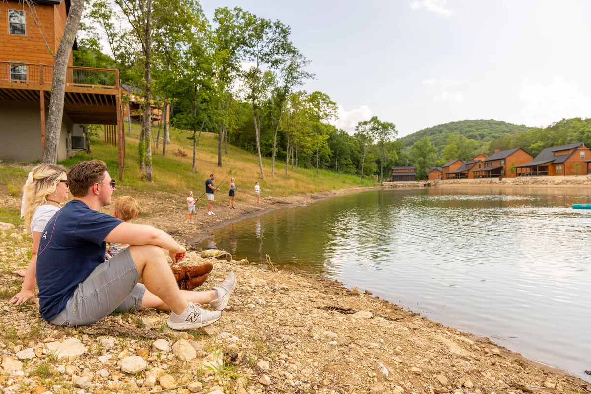 A tranquil lakeside scene with a family sitting by the water, children playing near the edge, and cabins and trees in the background. The mood is peaceful and inviting, encouraging fishing, water activities, and moments of connection with nature.