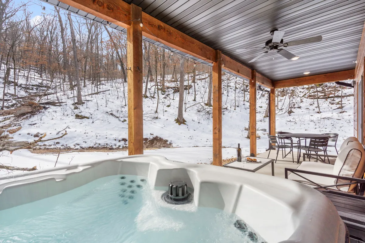 An inviting hot tub sits on a wooden deck framed by sturdy wooden pillars. The landscape is lightly dusted with snow, emphasizing year-round enjoyment and the magical ambiance of soaking outdoors in any season. Comfortable seating nearby suggests space for quiet moments together.