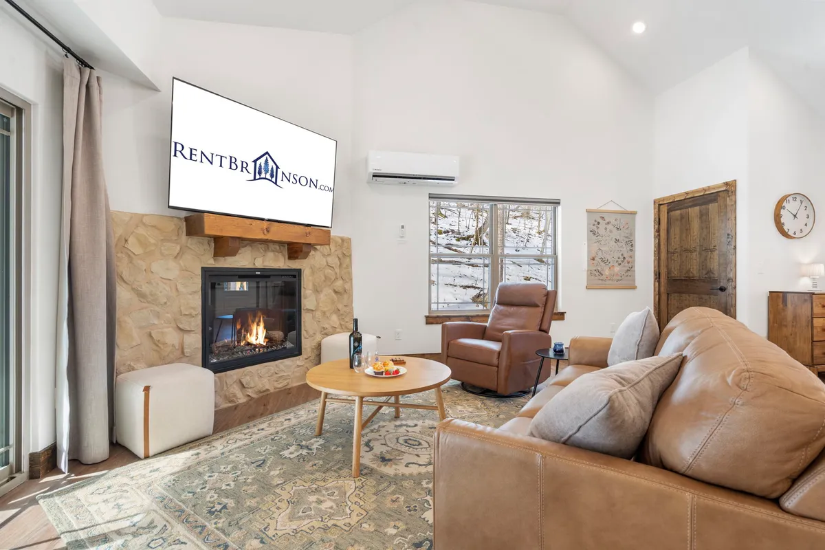 A welcoming living area featuring a fireplace as the centerpiece, a comfortable seating arrangement with a sofa and armchair, a large television, and a decorative rug. The space is bathed in soft, warm lighting, creating an inviting and relaxing atmosphere ideal for unwinding or spending quality time together.