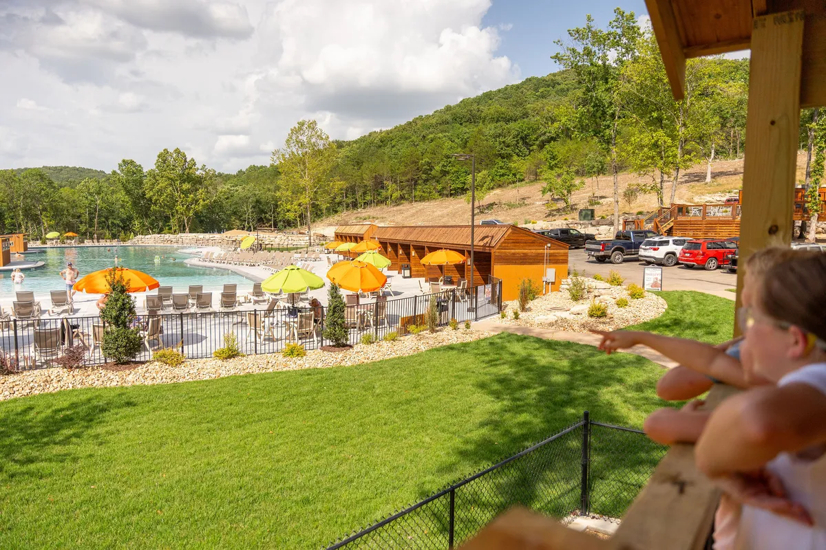 This exterior image captures the vibrant outdoor pool area at Tall Timbers Camp, featuring children playing in the pool, lounge chairs arranged around the water, colorful umbrellas, wooden cabins in the background, and a lush green lawn. The scene radiates lively group activity and summer fun.