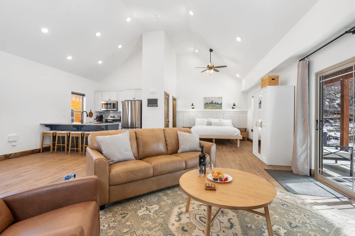 A bright, open-concept studio space with a sofa, coffee table, bed, kitchen area, and a sliding door letting in abundant natural light. This interior seamlessly connects living and sleeping areas, showcasing the flexibility and togetherness of the open-plan design. Sunlight highlights the wood accents throughout the space, enhancing the inviting mood.